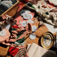 A styled flat-lay of vintage sewing and crafting supplies on a wooden table, including pink and red fabrics, white lace, circular blue-and-white patterned ornaments, a wooden frame, scissors, and papers under warm sunlight.