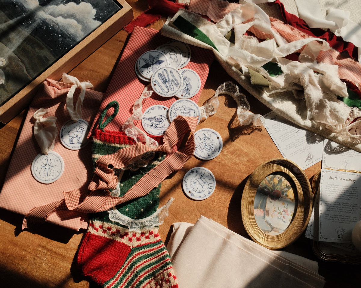 A styled flat-lay of vintage sewing and crafting supplies on a wooden table, including pink and red fabrics, white lace, circular blue-and-white patterned ornaments, a wooden frame, scissors, and papers under warm sunlight.
