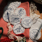 A styled flat-lay of circular white ceramic ornaments with blue illustrations, arranged on a red gingham cloth alongside lace ribbon, a green knit loop, and a printed card on a wooden surface.