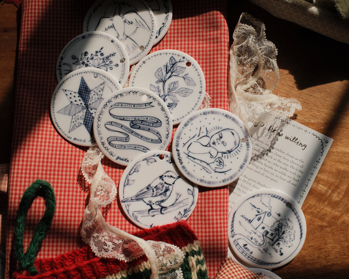 A styled flat-lay of circular white ceramic ornaments with blue illustrations, arranged on a red gingham cloth alongside lace ribbon, a green knit loop, and a printed card on a wooden surface.
