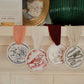 Four round decorative ceramic ornaments with illustrated designs hang from a white mantel with colorful ribbons. The ornaments feature blue, red, brown, and green line drawings, including a bird, a house, a lamb, and a ribbon.