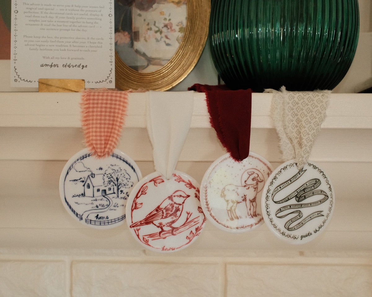 Four round decorative ceramic ornaments with illustrated designs hang from a white mantel with colorful ribbons. The ornaments feature blue, red, brown, and green line drawings, including a bird, a house, a lamb, and a ribbon.